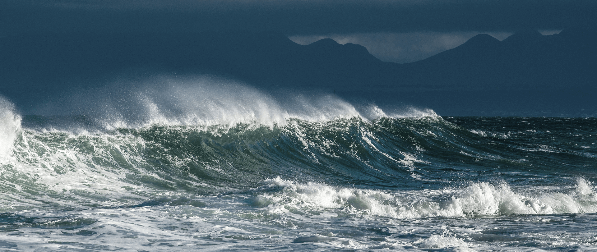 A wind swept ocean wave with sea spray flying off of the peak of the wave. Mountains silhouette the background.